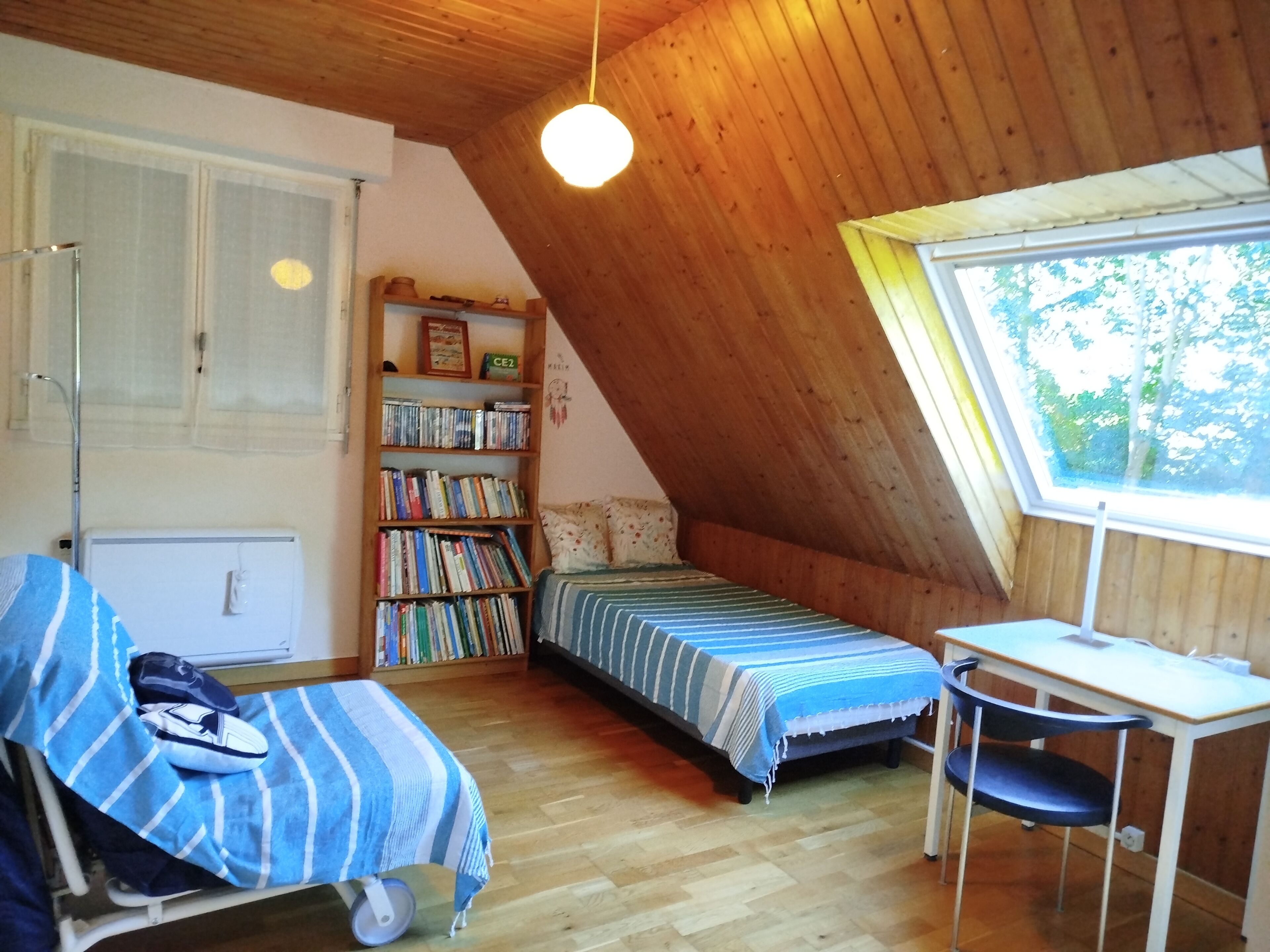 Une chambre mansardée chaleureuse, avec un plafond en pente, une bibliothèque en bois, un petit bureau et une fenêtre donnant sur les arbres.