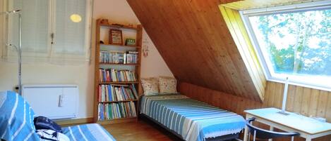 Une chambre mansardée chaleureuse, avec un plafond en pente, une bibliothèque en bois, un petit bureau et une fenêtre donnant sur les arbres.