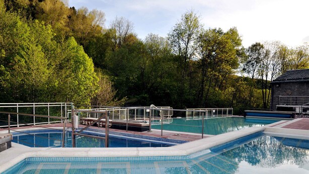 En swimmingpool med flisebelagt område og gelænder, omgivet af træer.