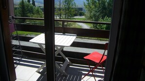 En balkon med et hvidt bord og to stole, en rød og en hvid, med udsigt over et smukt landskab med bjerge og grøn natur.