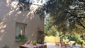 Une terrasse extérieure avec une table et des chaises, entourée de plantes et d’arbres.