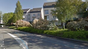 A residential street with apartment buildings, a clear sky, and well-maintained hedges.