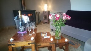 Un salon chaleureux avec une cheminée, une table basse en bois sur laquelle se trouve une bouteille de champagne, deux verres et un vase rempli de fleurs.