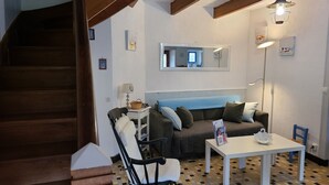 Ein gemütliches Wohnzimmer mit einer Couch, einem Esstisch und einem Sessel. Eine Treppe ist links zu sehen, und ein Spiegel hängt an der Wand.