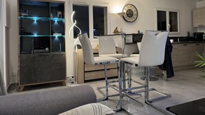 A modern living room with a sofa, two white chairs, a glass coffee table, and a kitchen area with cabinets and a clock. - CHARMING VILLA WITH PRIVATE POOL 4 TO 5 PEOPLE NEAR PALOMBAGGIA (PORTO VECCHIO)