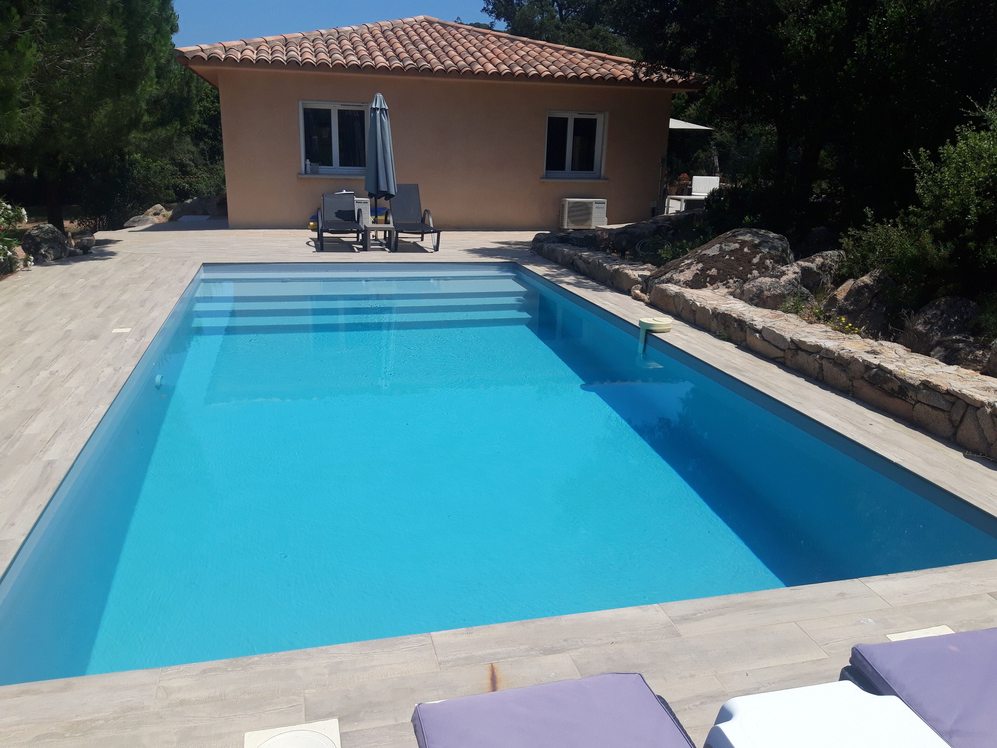 Une piscine rectangulaire bordée de pierres, deux chaises longues et, au fond, un petit bâtiment avec un toit en tuiles.