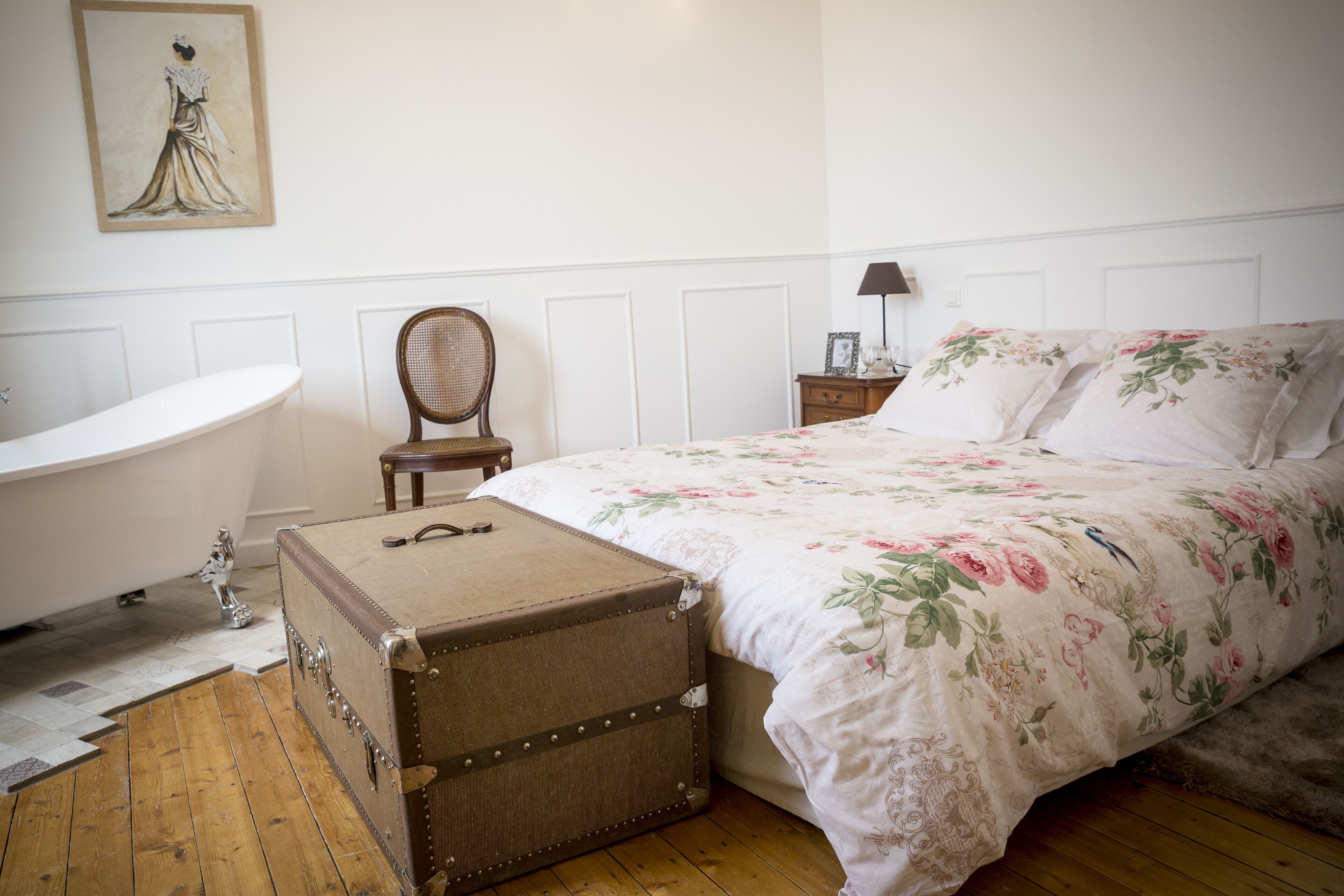 A bedroom with a large bed, a wooden floor, a vintage suitcase, a clawfoot bathtub, and a chair.