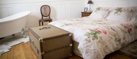 A bedroom with a large bed, a wooden floor, a vintage suitcase, a clawfoot bathtub, and a chair.