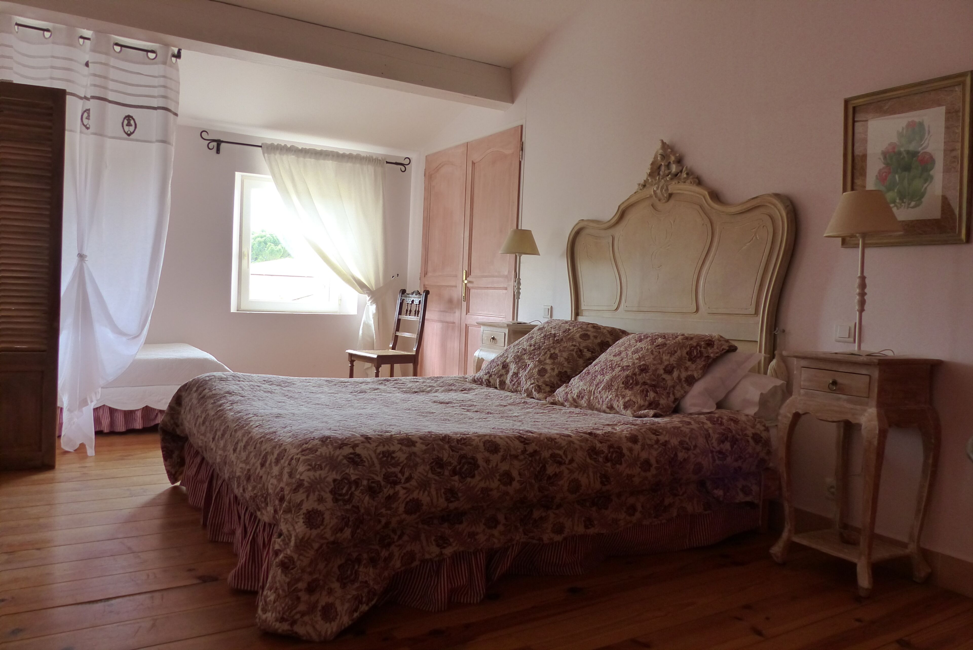 Una camera da letto con un letto grande, pavimento in legno, una finestra con tende, un comodino e un tavolino di fianco al letto.