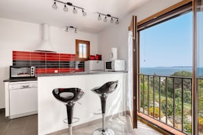 Eine moderne Küche mit roter und schwarzer Spritzschutzwand, einer weißen Kücheninsel mit zwei Barhocker und Blick auf das Meer vom Balkon aus.