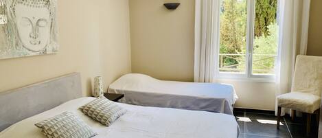 A room with two beds, a chair, a window with curtains, and a wall-mounted light fixture.