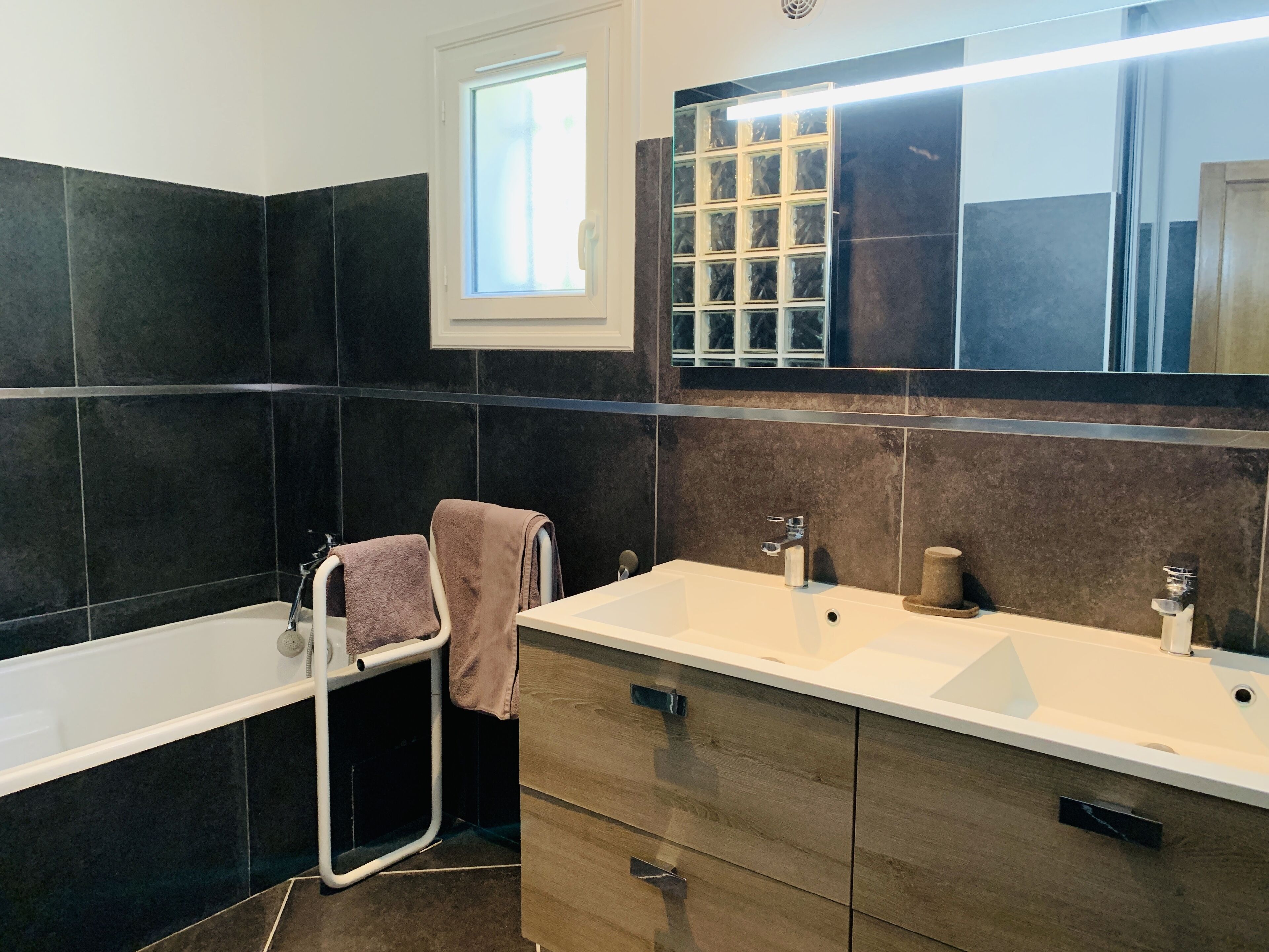 A modern bathroom with dark tiles, a large mirror, a window, and a bathtub.