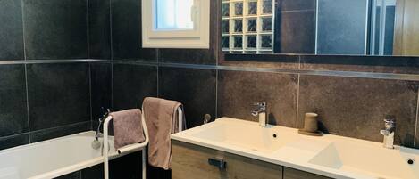 A modern bathroom with dark tiles, a large mirror, a window, and a bathtub.