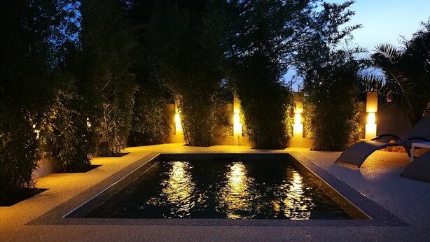 A rectangular pool with a surrounding paved area, illuminated by warm lights, surrounded by dense foliage and a few lounge chairs.