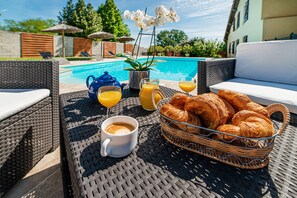 A wicker table with a basket of croissants, a cup of coffee, and a glass of orange juice by a poolside.