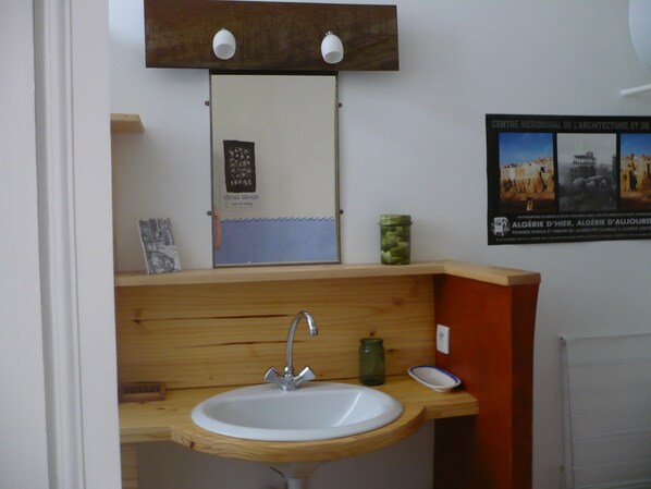 A bathroom with a wooden vanity, a white sink, and a mirror above it. There is a small shelf with a framed picture and a green jar.