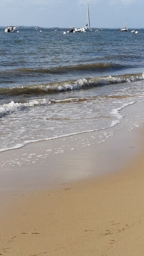Une plage où les vagues viennent doucement lécher le rivage, un ciel dégagé et plusieurs bateaux visibles au loin.