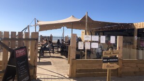 Una estructura de madera frente a la playa con mesas y sillas, un cartel que dice '¡Bienvenidos!' y 'Bar & Snacking', y una vista de la playa al fondo.