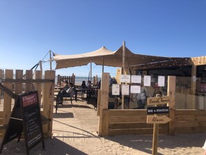 Una struttura in legno sul lungomare con tavoli e sedie, un cartello con le scritte 'Welcome!' e 'Bar & Snacking', e sullo sfondo la spiaggia.