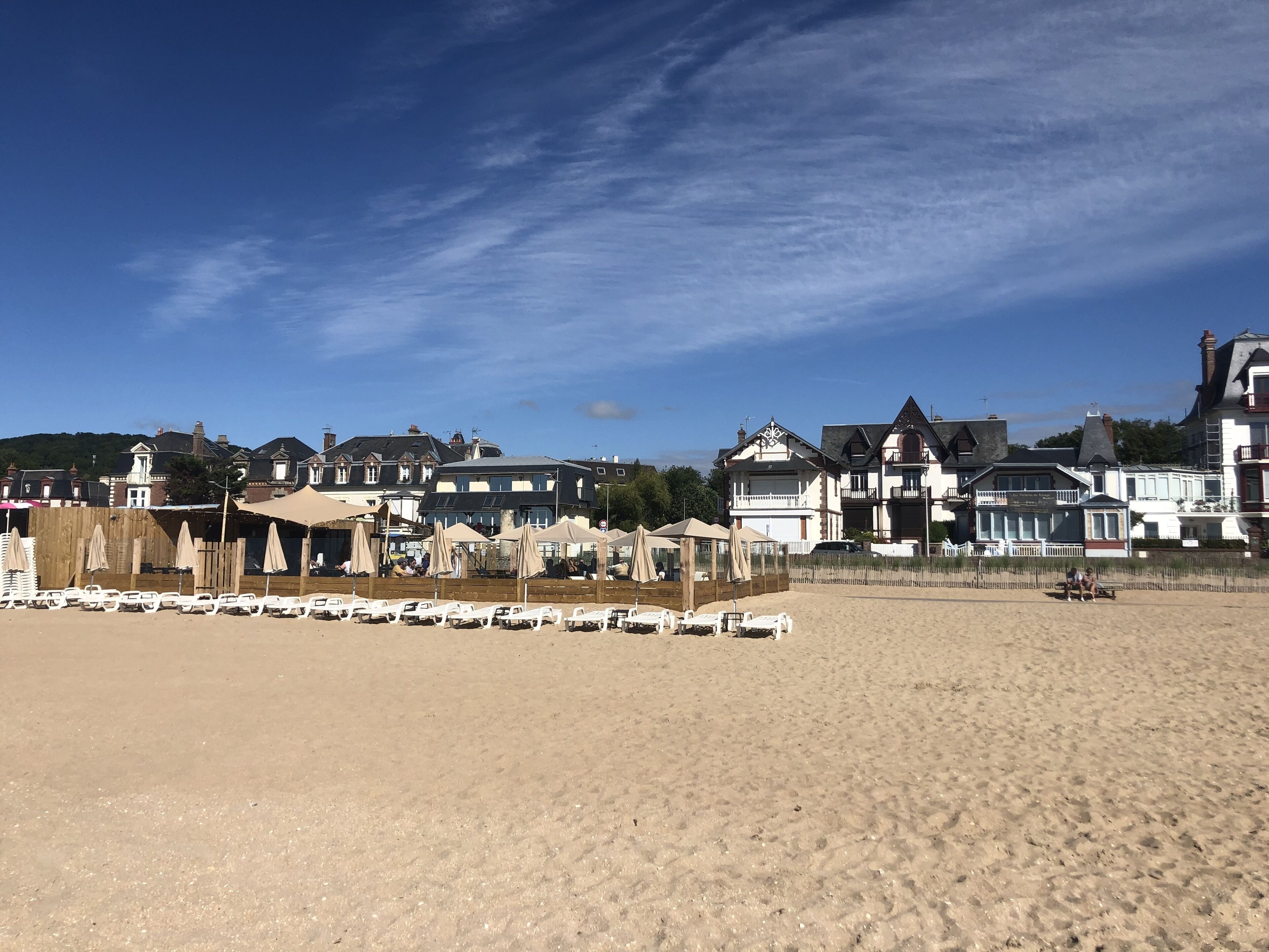 Ein Strand mit Reihen aus weißen Liegestühlen und Sonnenschirmen, eine Holzlattenveranda und eine Reihe großer, historischer Häuser im Hintergrund.