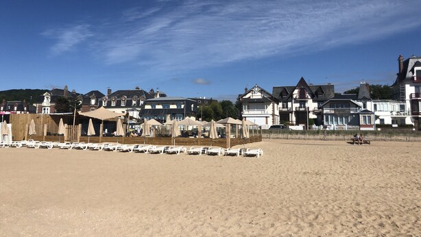 Una playa con hileras de sillas de playa blancas y sombrillas, una cerca de madera y una fila de casas grandes e históricas al fondo.