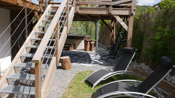 Eine Holzterrasse mit einer Treppe zu einer erhöhten Ebene, Sitzgelegenheiten im Freien mit Liegestühlen und einem kleinen Tisch.