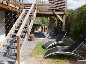 Une terrasse en bois avec un escalier menant à un niveau supérieur, un espace extérieur aménagé avec des fauteuils et une petite table.