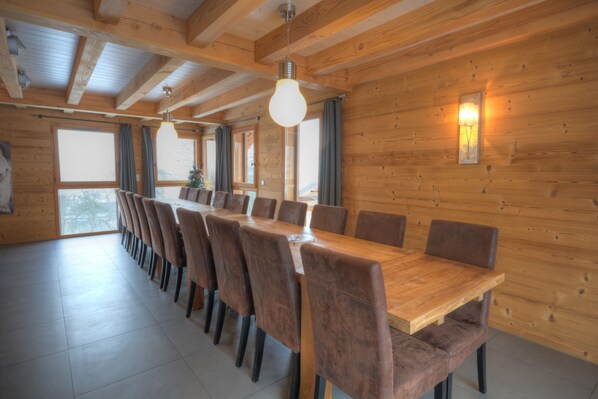 A wooden-paneled dining area with a long table and chairs, large windows, and a hanging light fixture. - Grand Massif 8 bedrooms/8 bathrooms sauna, pool table, walk to village & gondola (Morillon)