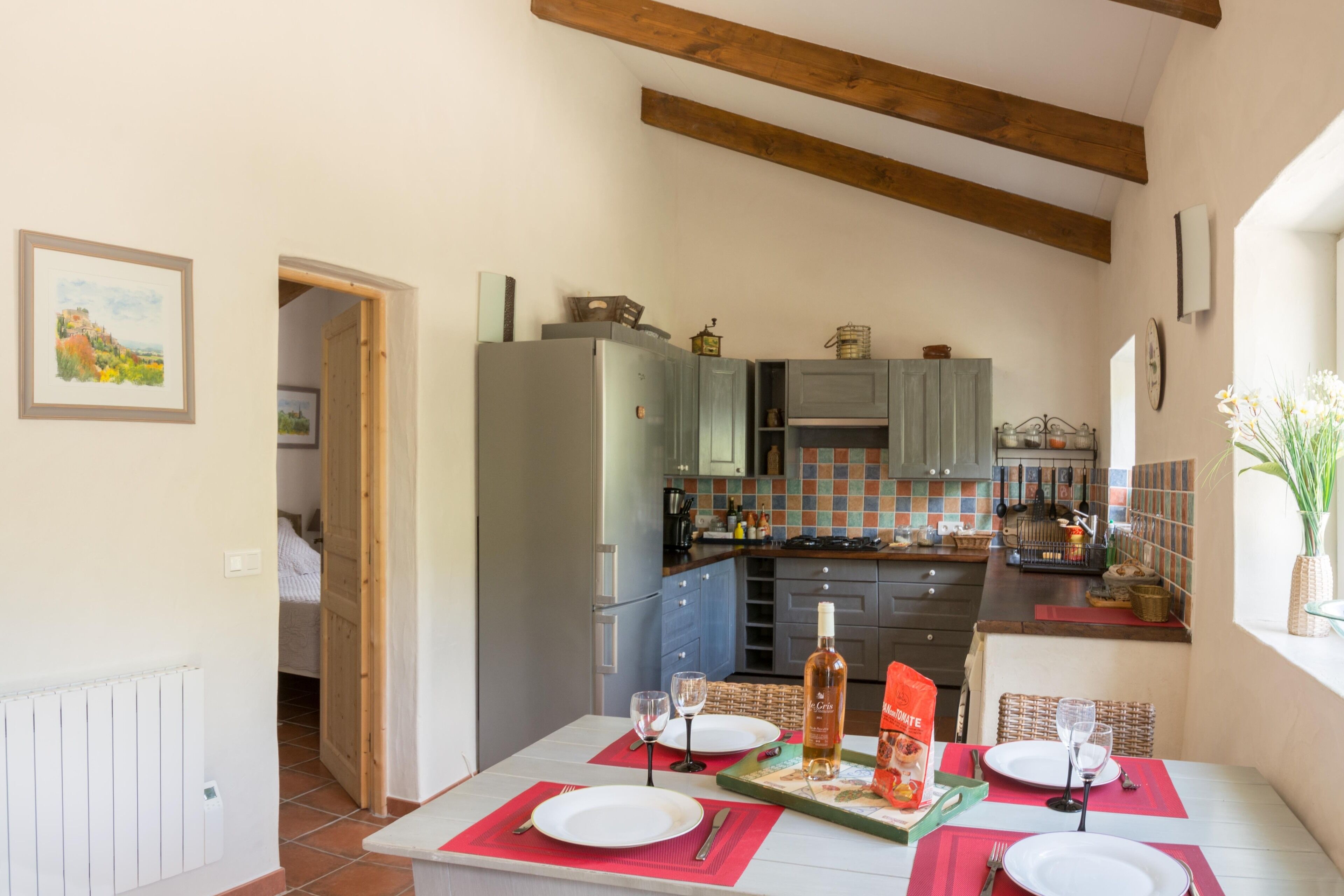 Un coin repas avec une table dressée pour deux personnes, une cuisine en arrière-plan et une vue sur la campagne par une porte ouverte.