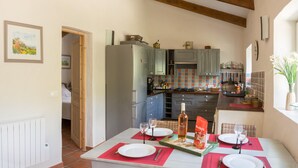 A dining area with a table set for two, a kitchen in the background, and a view of the countryside through an open door.