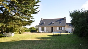 Une maison en pierre avec un toit en pente, un petit porche à l’avant et une pelouse aménagée avec du mobilier d’extérieur.
