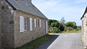 Un bâtiment en pierre avec un toit en pente et des volets blancs, une route étroite qui y mène, et un panneau « interdiction de stationner » visible au loin.