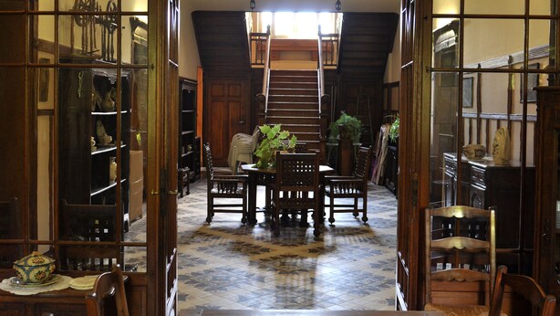 Un couloir avec un escalier en bois, un coin repas avec une table et des chaises, et des étagères remplies de divers objets.