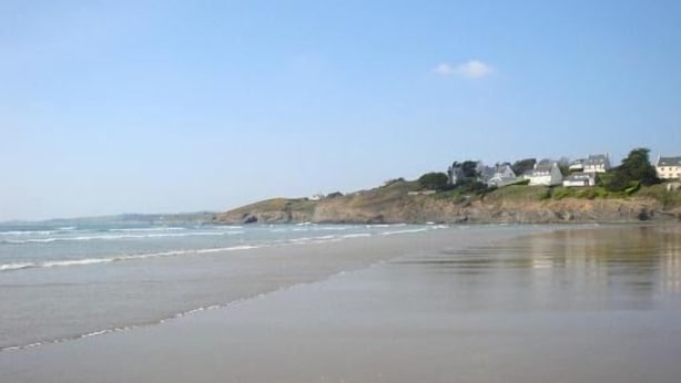 Une plage avec un ciel dégagé, une mer calme et des maisons situées sur une falaise.