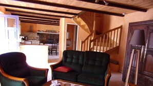 Un salon chaleureux avec un canapé à motifs verts, une table basse en bois et une chaise en bois. On aperçoit un escalier au fond de la pièce, menant à un niveau supérieur.