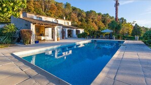 A rectangular swimming pool with a tiled deck, surrounded by a house with a tiled roof and stone walls, set in a landscaped garden with trees and a palm tree. - Starry EDEN (Saint-Paul-De-Vence)