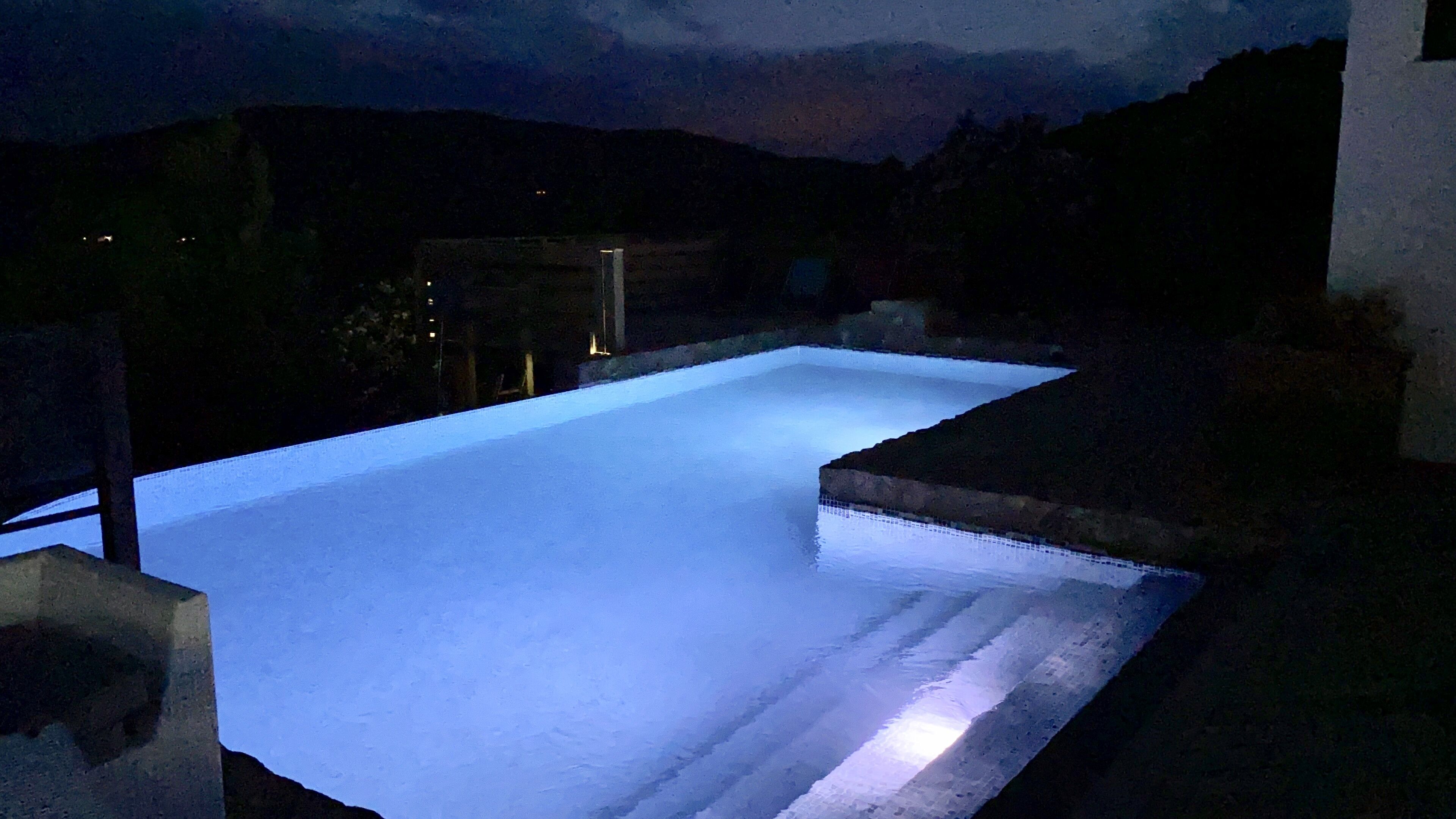 Una piscina rectangular con borde de piedra, rodeada por un muro bajo y un paisaje oscuro al fondo.