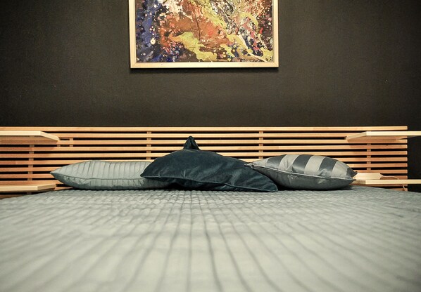 A bed with a quilted grey bedspread and two pillows, a wooden headboard, and a colorful abstract painting on the wall.