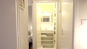 Ein schmaler Korridor mit Holzboden, weißen Wänden und einer Deckenleuchte. Eine Tür rechts führt zu einem Badezimmer mit Spiegel, Waschbecken und Handtuchhalter.