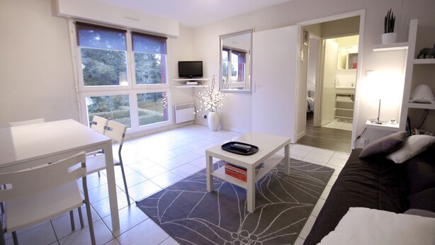 Ein modernes Wohnzimmer mit einem weißen Esstisch, einer Couch und einem Fernseher. Ein großes Fenster lässt Tageslicht herein und bietet Blick auf Bäume.