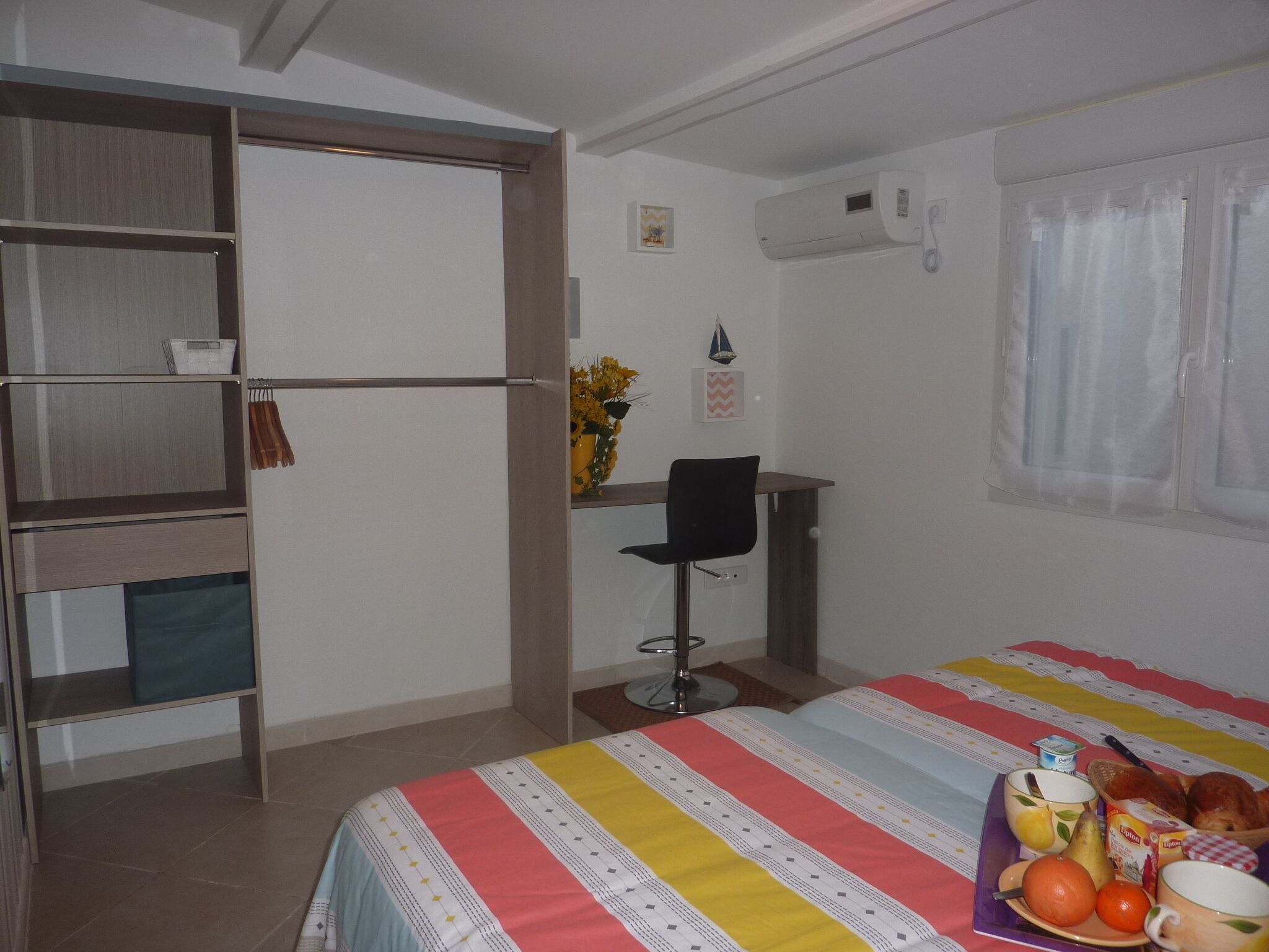 Un dormitorio con cama, escritorio y silla. Hay una ventana con cortinas, aire acondicionado de pared y una estantería con una cesta y un jarrón con flores.