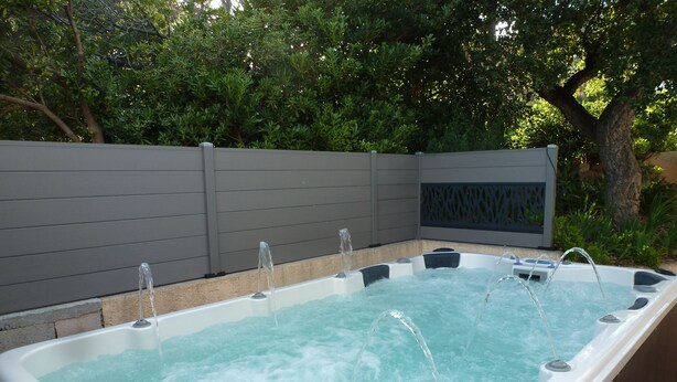 Ein Whirlpool mit mehreren Düsen, umgeben von einer Holzterrasse und einem grauen Zaun.