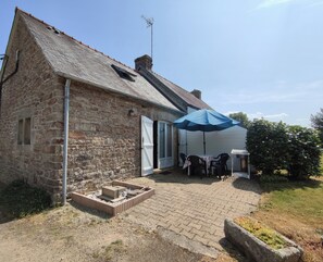 A stone building with a tiled roof, a patio area with a table and chairs, and a blue umbrella.