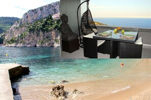 A serene coastal scene with a Buddha statue overlooking a clear blue sea, a dining area with a hanging chair, and a rocky cliff in the background.