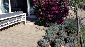 Una terraza de madera con dos jarrones grandes, un edificio blanco de techo con tejas y un arbusto morado con flores.