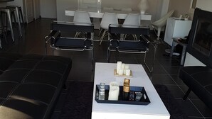 Una sala moderna con un sofá de cuero negro, una mesa de centro blanca con velas y un televisor. Se ven sillas blancas y una zona de bar al fondo.
