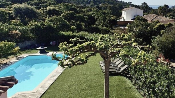Una piscina rodeada de un césped cuidado, una pérgola con enredaderas y vistas a una colina con edificios.
