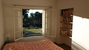 Um quarto com janela aberta, cama com colcha estampada, abajur e estante de livros.