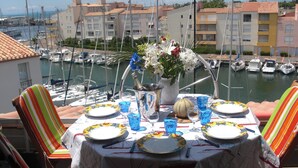 A dining table set with plates, glasses, and a vase of flowers, overlooking a marina with boats and buildings. - Grand Confort two-bedroom apartment with sea view and channel Near Sea (Agde)