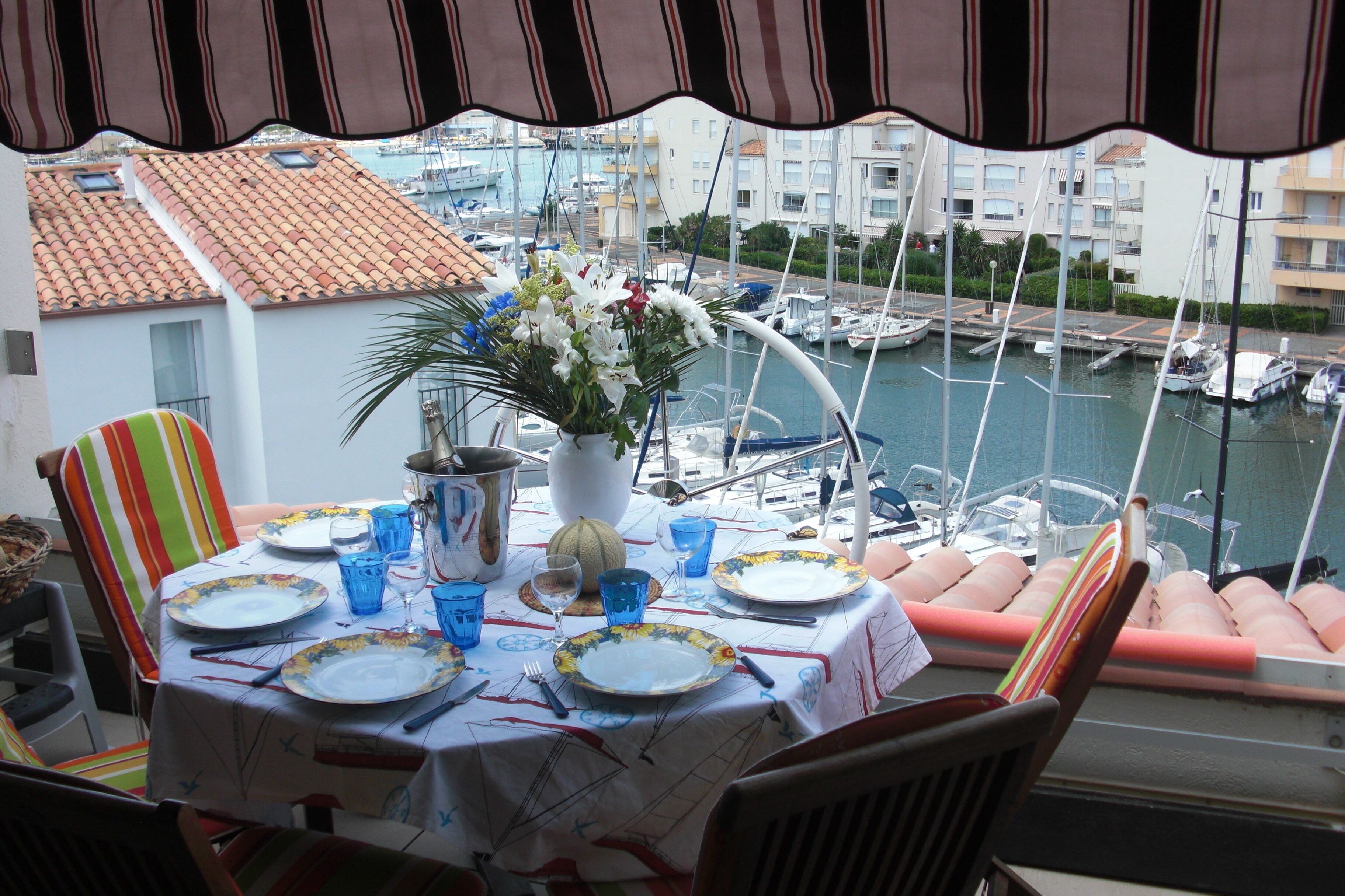 A dining table set with plates, glasses, and a vase of flowers, overlooking a marina with boats and buildings.